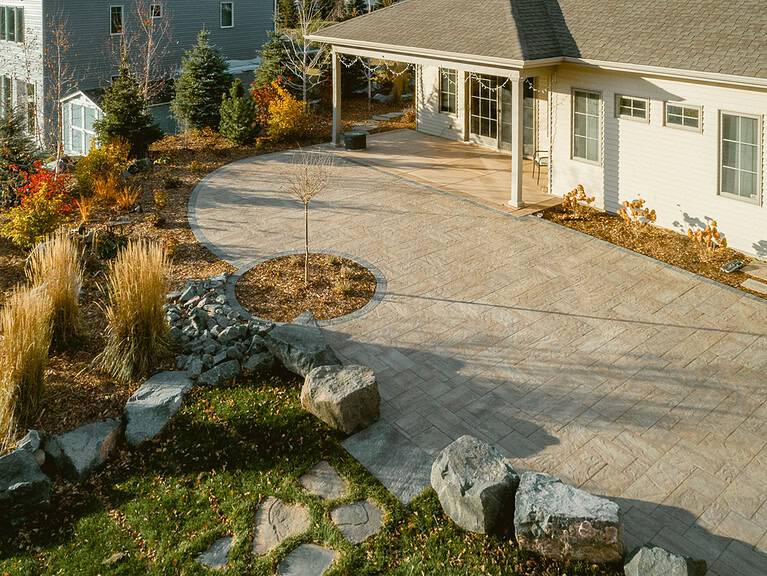 Stamped concrete patio with a circular tree inset, boulder edging, and ornamental grasses by Miller Creek